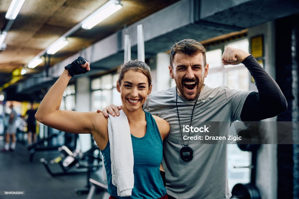 Femme athlétique heureuse et son professeur de fitness contractant leurs bras dans une salle de sport tout en regardant la caméra.